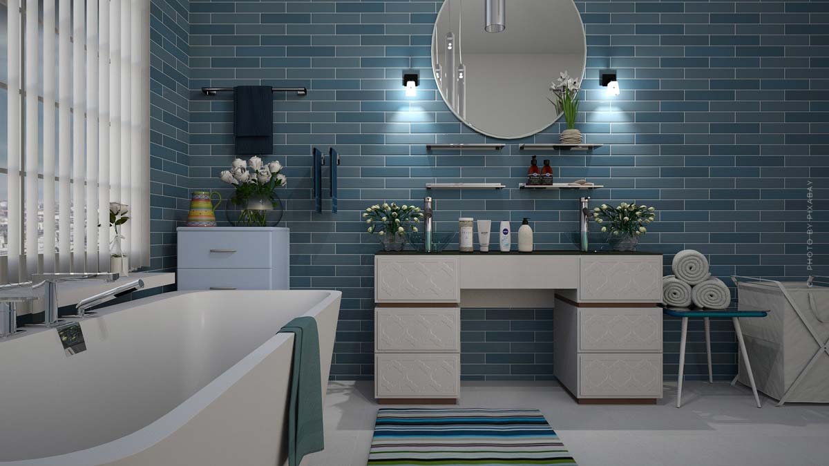 bad-badezimmer-blaue fliesen-himmelsfarben-metalltöne-offene badewanne-waschtisch-spiegel-sauber-handtücher-blumen-teppich gestreift