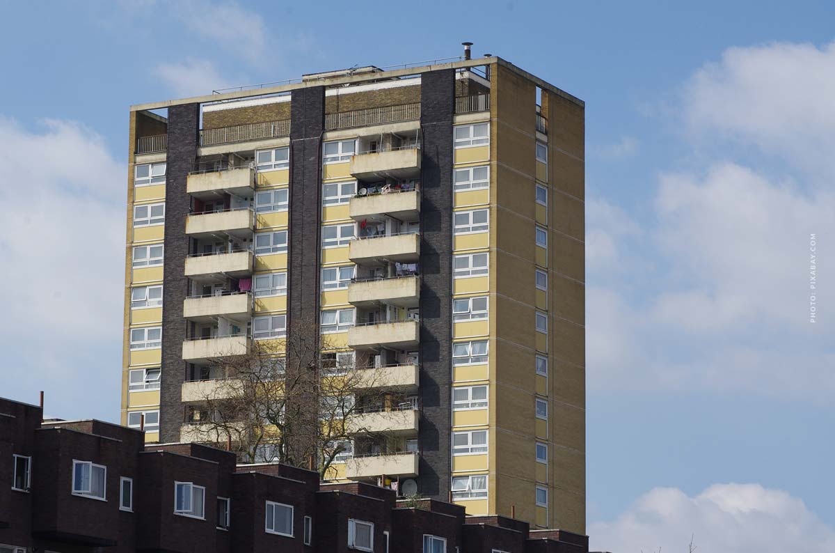 Hier sehen Sie ein Hochhaus mit gelber Fassade und mehreren Balkonen an einem Stadtrand. 